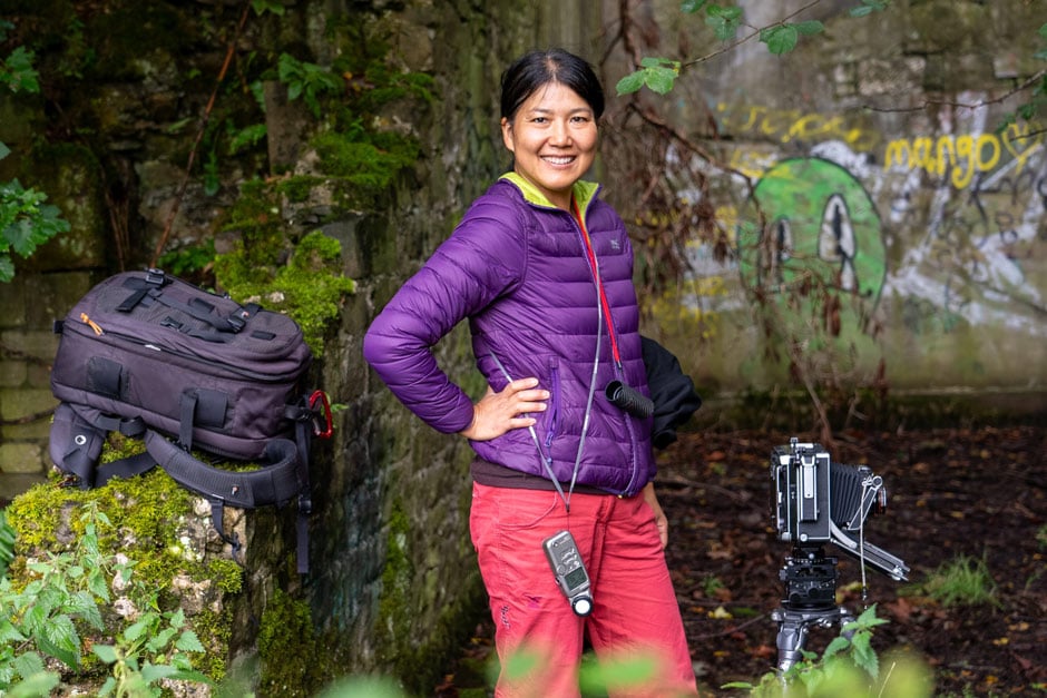 Dr Yan Wang Preston stands confidently in bright colourful clothing, with a historical large format camera in front of a mossy wall at RHS Bridgewater.