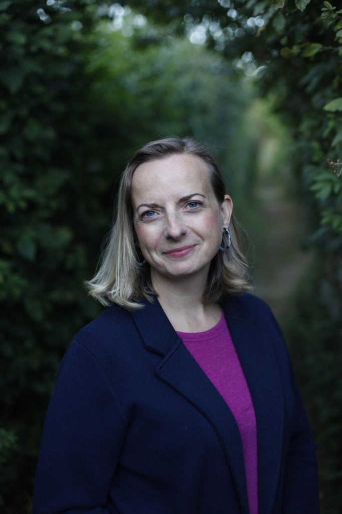 A portrait of Liz Lock, a white woman with blond shoulder length hair, stood in fronts of trees in soft focus.