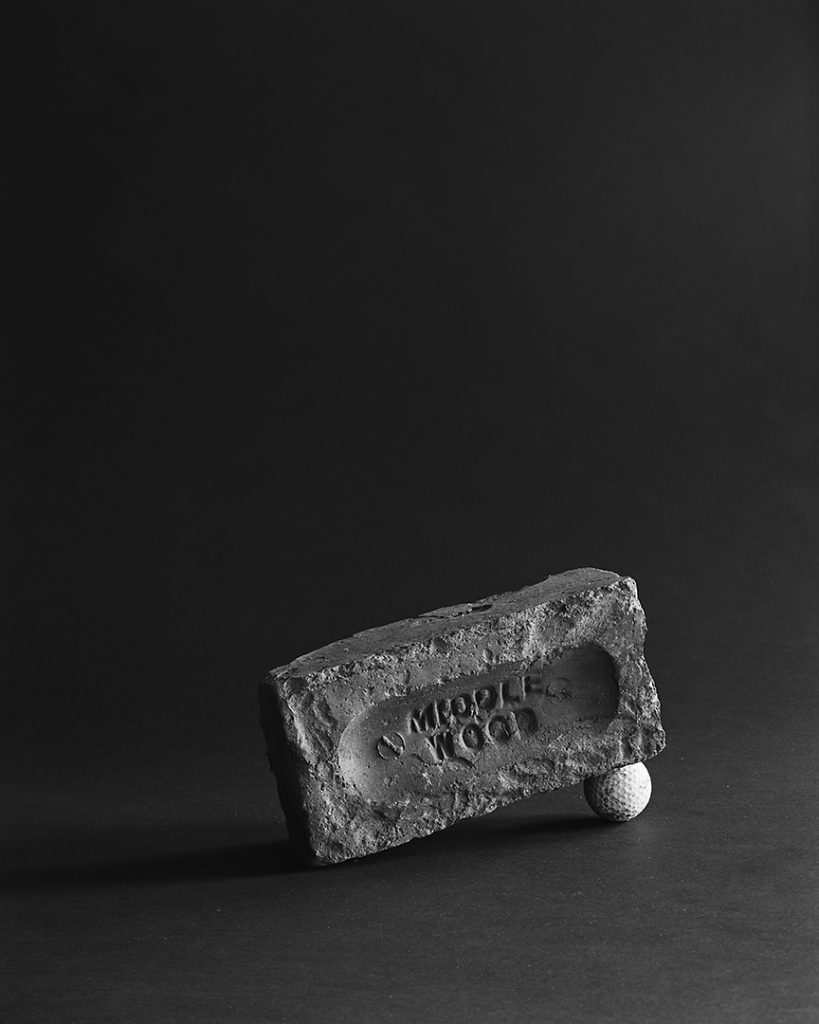A bricked marked with the name 'Middle Wood' is photographed in black and white, it is tilted on a ping pong ball and is photographed against a black background with side lighting.